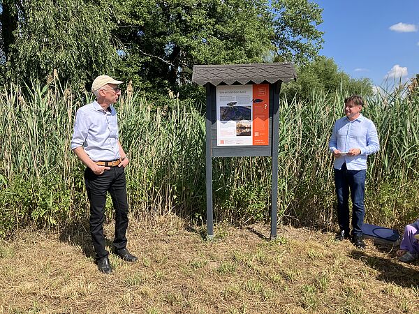 Zwei Männer stehen neben einer Infotafel am Wegesrand.