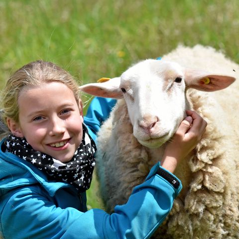 Mädchen mit Schaf auf grüner Wiese schaut lächelnd in die Kamera 