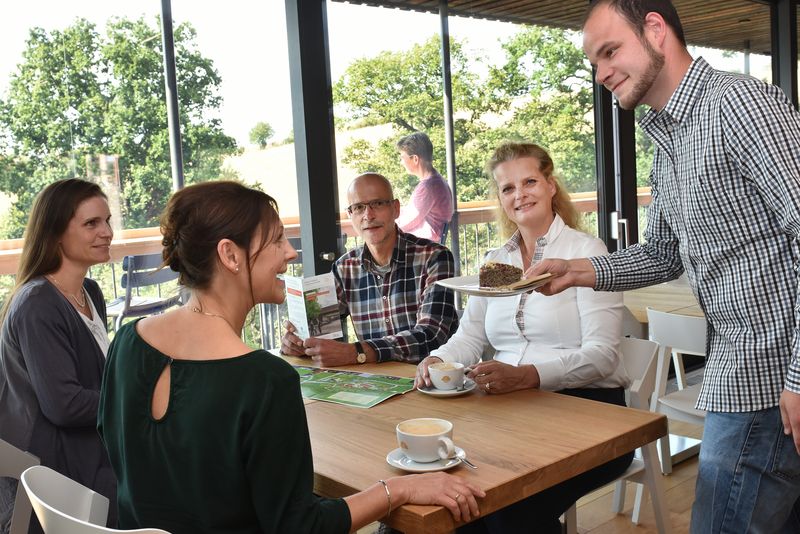 Café GUT(e) auszeit: Vier Personen am Tisch vor Panoramafenster, Kellner bringt Kuchenteller