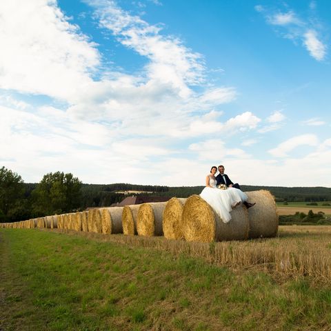 Hochzeitspaar sitzt auf Heuballen am Feldrand vor blauem Himmel
