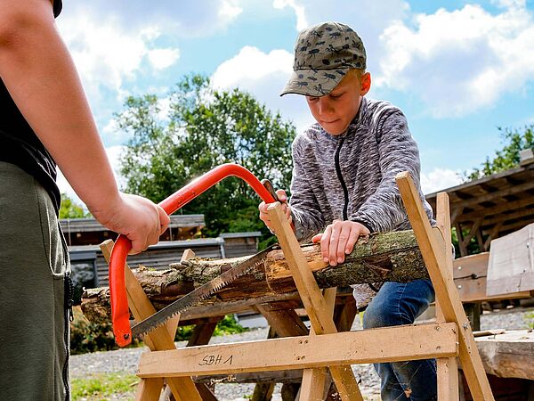 Mitmachen ausdrücklich erwünscht: Sommerferienprogramm auf Gut Herbigshagen Junge mit Basecap sägt unter Anleitung konzentriert ein Holzstück durch