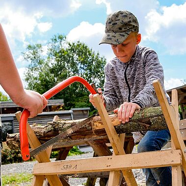 Junge mit Basecap sägt unter Anleitung konzentriert ein Holzstück durch