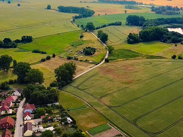 Intakte Biotopverbünde tragen nicht nur zum Erhalt der Biodiversität bei, sie haben auch viele positive Effekte auf die Regionen. Aufsicht auf Häser und Straßen einer kleinen Ortschaft. Großteil des Bildes ist von landwirtschaftlichen Feldern ausgefüllt. Ein mit Bäumen und Büschen gesäumter See ist am rechten Bildrand zu sehen.gesäumter