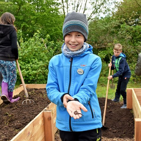 Junge in blauer Jacke zeigt stolz einen Regenwurm auf seiner ausgestreckten Hand, mehrere Kinder arbeiten an Hochbeeten im Hintergrund