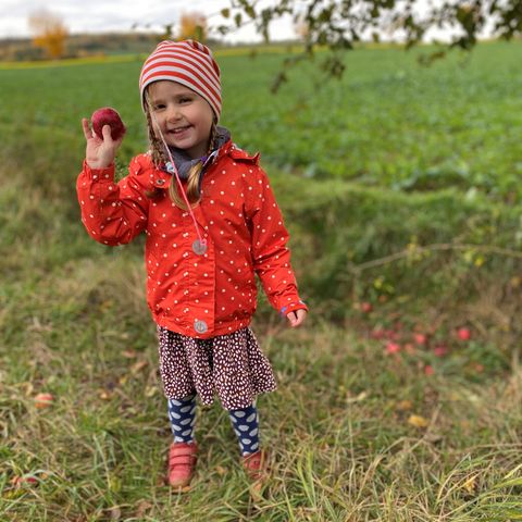 Kind mit roter Jacke und Mütze steht auf Wiese und hält Apfel in der Hand