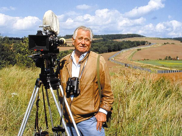 Heinz Sielmann bei Dreharbeiten zu "Tiere im Schatten der Grenze", 1988 Heinz Sielmann mit Kamera und Fernglas vor einer Landschaft