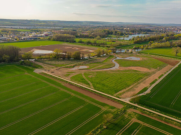 Die Biotoplandschaft am Flüthewehr in Göttingen nach den umgesetzten Maßnahmen. Aus der Vogelperspektive sind die neuen Strukturen auf den Flächen zu erkennen. Dazu zählt ein Weiher und mehrere temporäre Tümpel. Links und rechts sind intensiv genutzte Ackerflächen. Am Horizont befinden sich einige Gebäude der Stadt Göttingen. Die Leine fließt durch dieses Areal.