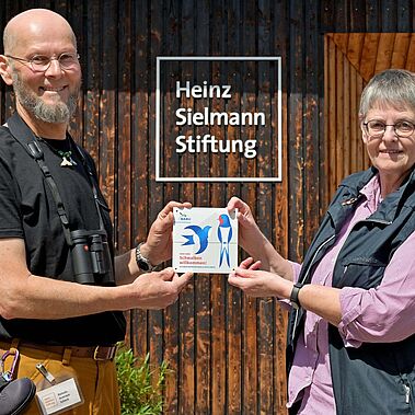 Zwei Personen stehen mit einer Auszeichnung für ein schwalbenfreundliches Haus in den Händen vor einer Holzwand
